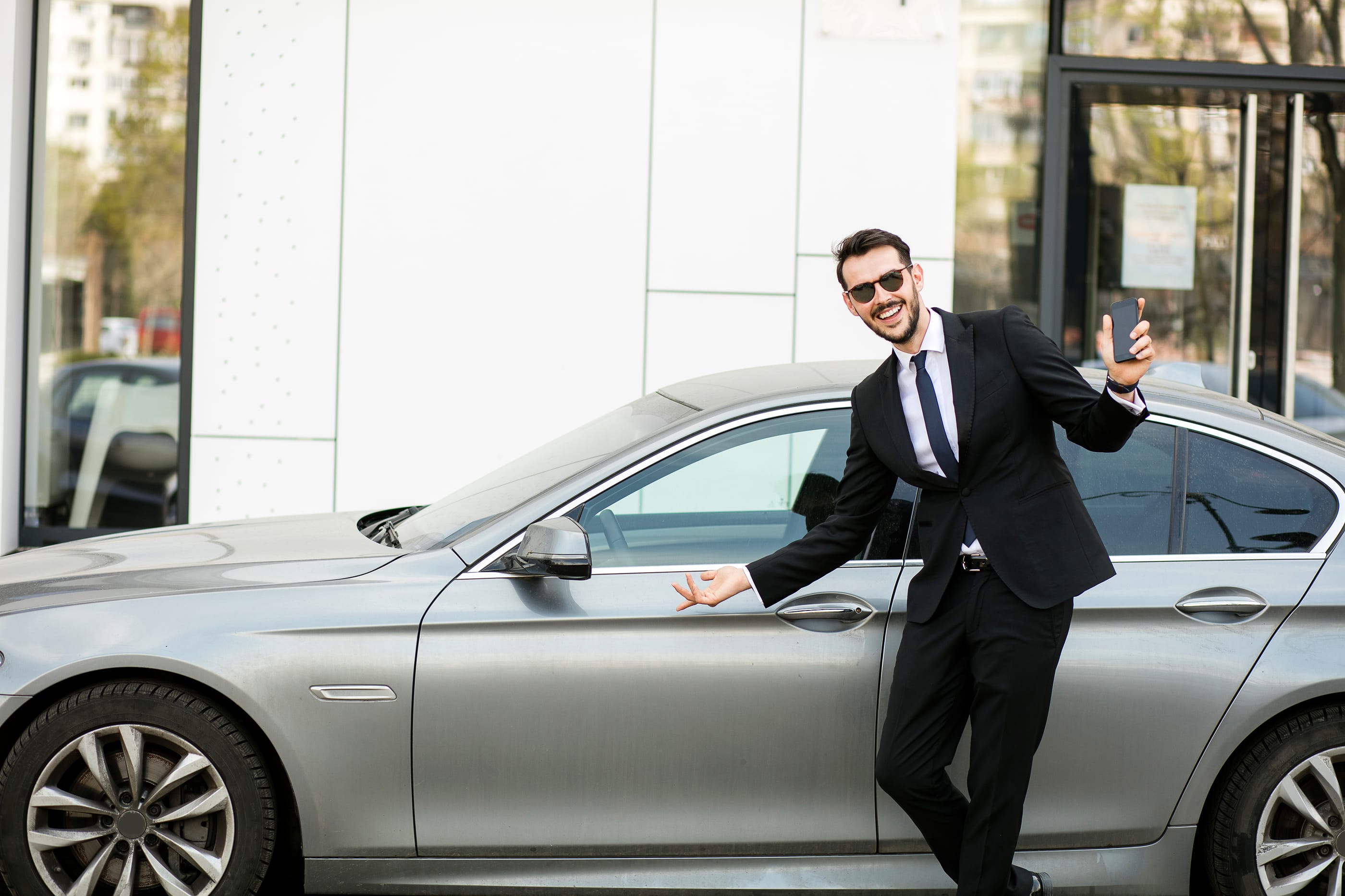 uber driver in elegant suit in front of an luxury car showing his cellphone, outside on the street in front of an office building